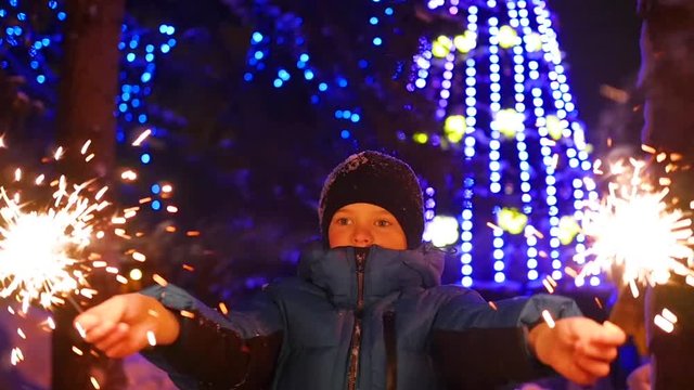 The child holds the sparklers outdoors in the winter. Slowmotion . In the background, lights and garlands of Christmas fir