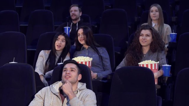 Young people watching movie at the movie theater. Two boys and four girls sitting on different rows at the cinema. Asian teenage girl talking to caucasian girl