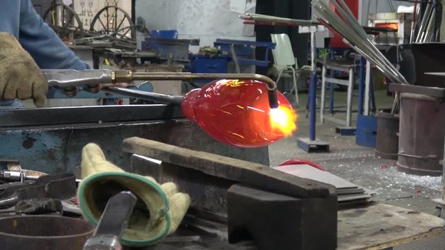 Footage of glassblowing workshop factory glassblower also known as glassmith or gaffer working on vase using a blow torch while turning glass crystal from oven is glowing yellow orange from heat 4k