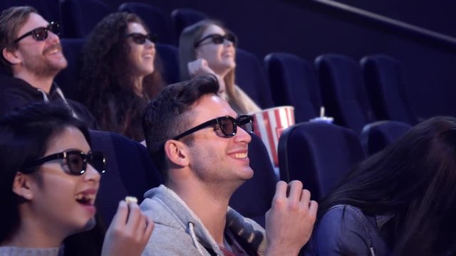 Young people in 3D viewers laughing at the movie theater. Caucasian brunette girl touching her hair at the cinema. Asian girl taking flake of popcorn from the bucket of caucasian guy