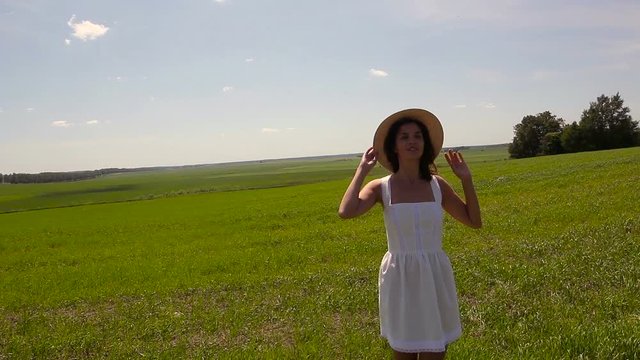 young girl stands on the site in white dress and clothes straw hat is on the green field with grass and smiles, holds the hand hat, summer. whirling around of the camera model