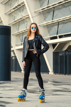 Girl on rollerblades standing in building background. Young fit women girl in blue sunglasses, jeans and jacket on roller skates riding outdoors after rain.
