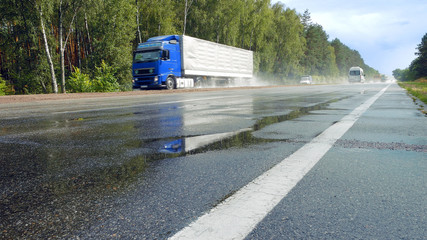Naklejka premium View from ground of traffic on wet road, highway with mist splash after rain with green trees on a roadside on background. Trucks are passing by camera in perspective
