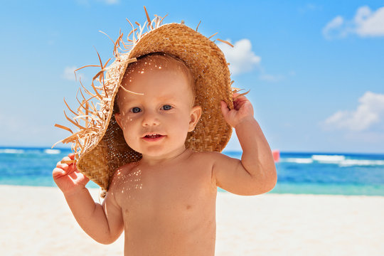 Funny photo of happy baby boy on beach with straw hat and dirty face covered with sand. Family travel, healthy lifestyle, recreation, water outdoor activity on summer beach vacation with children.