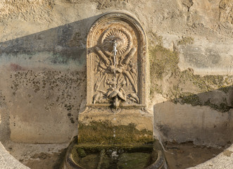 Ancient water fountain carved in stone, with restored modern faucet water falling, side illuminated by the sun