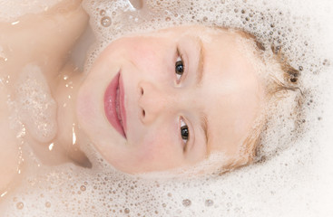 Fototapeta premium Cute ginger haired boy lying in the bath which is fullof bubbles. His head is prtly submerged and he is looking into the camera. Upper body shot includes head and shoulders.