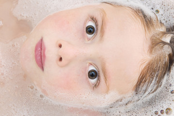 Naklejka premium Close up shot of cute ginger haired boy lying in the bath. He is looking directly into the camera and his head is partly submerged in the water which has bubbles in it. 