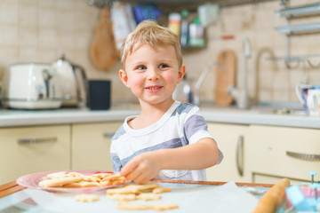 Little blond kid tries freshly baked shortbread, sitting at the kitchen table. Teeth smiling face.