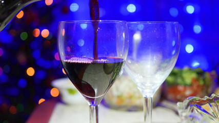 Red wine is poured into glasses. In the background, bokeh lights and garlands of Christmas fir