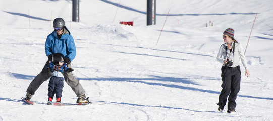 Toddler Learns to Ski with Dad While Watches & Takes Photos. Safely Dressed with Helmet, Sunglasses...