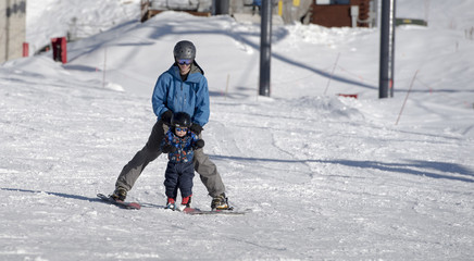 Fototapeta na wymiar Toddler Learning to Ski with his Father at a Colorado Resort. Dressed Safely with Helmet, Sunglasses, & Harness.