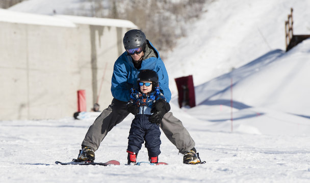 Toddler Learns to Ski with Dad. Safely Dressed with Helmet, Sunglasses, & Harness