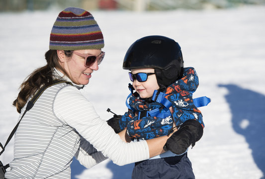 Toddler Learns to Ski at a Colorado Resort With Mom. Dressed Safely with Helmet, Sunglasses & Harness