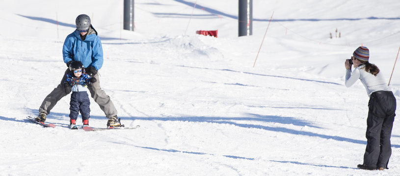 Toddler Learns to Ski with Dad While Mom Takes a Photo. Dressed Safely with Helmet, Sunglasses & Harness