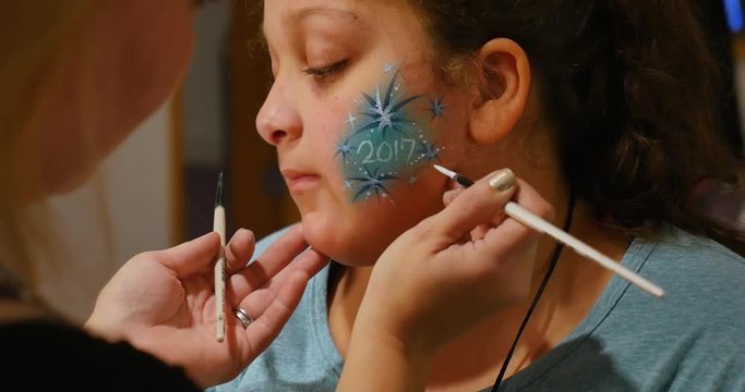 A little girl gets her face painted at a carnival or craft show.	 	