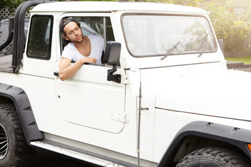 People, lifestyle and travel. Stylish young man driving his white four-wheel drive vehicle, sticking his head out of open window and looking ahead at road on safari trip in wild nature on sunny day © WHstudio Leushin N