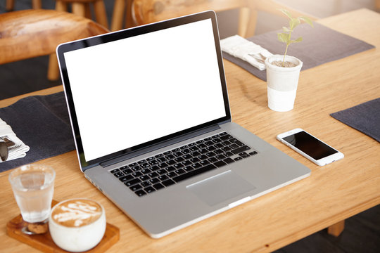 Business, technology and communication concept. Minimalistic workspace with modern laptop computer with white blank screen, generic mobile phone, coffee and glass of water on wooden desk. Mock up