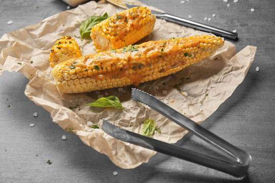 Tasty grilled corncobs with basil, sauce and tongs on grey table, close up view