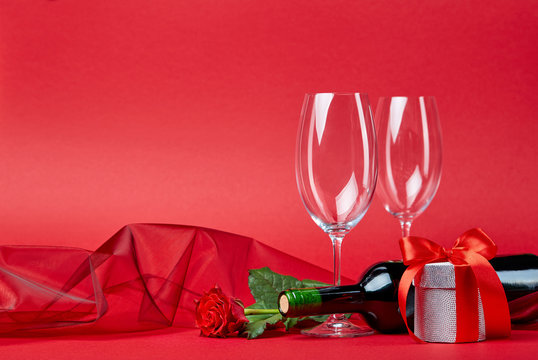 Red background with gift, bottle of wine, glasses and rose