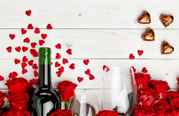 Buds of red roses and bottle of wine on white wooden background with confetti, glass and chocolate
