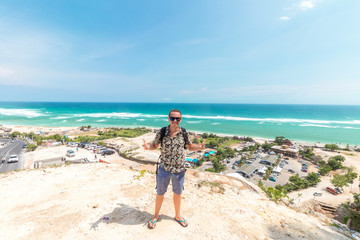 Handsome traveler man stay by blue ocean background - Happy guy relaxing at sea view point - Concept of freedom and summer trip around the world backpacker style. Bali, Indonesia.