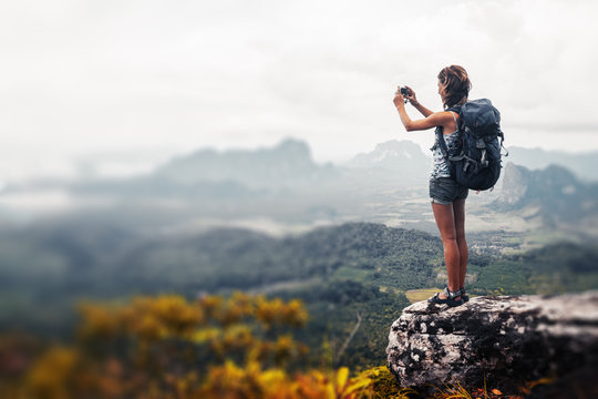 Young lady hiker with backpack standing on top of the mountain and taking a picture of valley