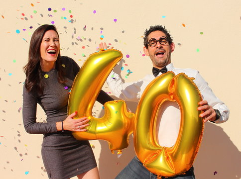 Cheerful couple celebrates a forty years birthday with big golden balloons and colorful little pieces of paper in the air.