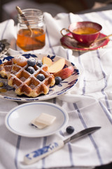 Blueberry apple waffle with honey set on a dish decorated with fresh blueberry, apple, tea pot set ready to serve.