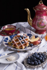 Blueberry apple waffle with honey set on a dish decorated with fresh blueberry, apple, tea pot set ready to serve.