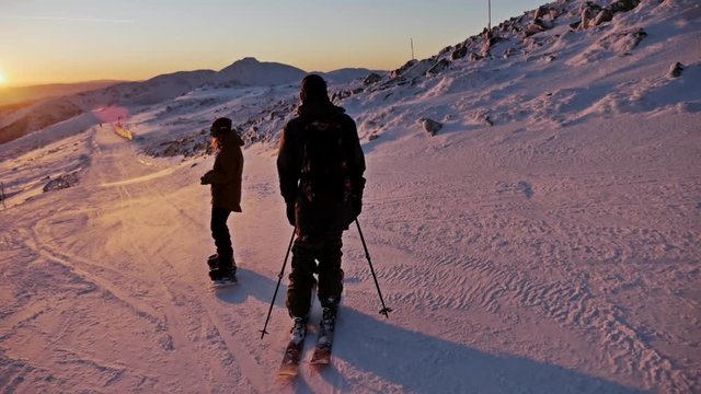 Slowmotion footage of a skier and snowboarder riding the slope during a beautiful sunset.