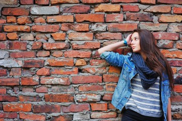 girl with long hair, dressed in a denim jacket and a scarf standing near a wall made of red bricks and waits