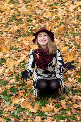 Fototapeta na wymiar Woman in a floral patterned coat and wine red hat playing with leaves in the park. Happy girl throws maple tree leaves into the air. Yellow, green, red and brown leaves, colorful autumn forest