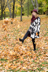 Woman in a floral patterned coat and wine red hat playing with leaves in the park. Happy girl throws maple tree leaves into the air. Yellow, green, red and brown leaves, colorful autumn forest