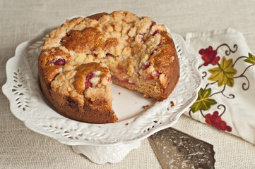 Apple crumb sour cream coffee cake on a round white elevated platform with a wedge cut out by a silver pie server with a colorful, patterned, cloth napkin