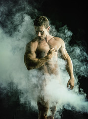 Totally naked male bodybuilder with smoke hiding genitalia, looking away to a side, on dark...