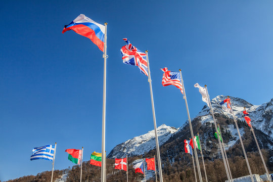 National flags of Russia, UK, USA, Greece and other countries wave in the wind as a friendship symbol on snowy mountains peak and blue sky background. Various fluttering national flags on flagpoles
