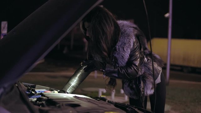 Young brunette woman with long hair is looking under the hood using her smartphone as a flashlight