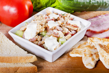 Simple, fresh salad with vegetables and meat in white bowl on a wooden board next to toast, cucumber, tomato, grilled chicken and smoked ham.
