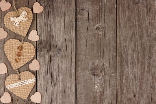 Side border of handmade burlap hearts with ribbon and buttons over a rustic wooden background