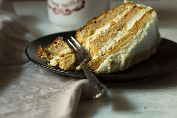 Slice of simple homemade carrot and honey cake with cream cheese frosting on a dark plate, broken off piece with fork, cup of coffee or tea, cozy atmosphere