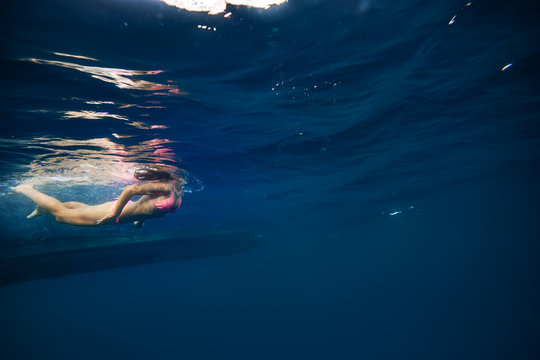 Girl in pink bikini underwater near bottom of a boat. Copy space on right side of image with blue water ocean background. Water surface with ripples above a model