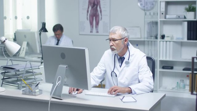 Close-up of a Gray Haired Senior Doctor and His Assistant Working at Desktop Computers. Sitting in Brightly Lit Office.  Shot on RED Cinema Camera in 4K (UHD).