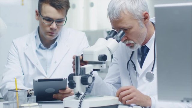 Senior Male Doctor Conducting Health Issue Investigation Through Microscope. His Young Assistant Writes all Relevant Data in Tablet Computer.Shot on RED Cinema Camera in 4K (UHD).