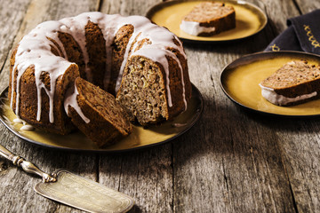 Banana cake with apples and walnuts drizzled frosting and sliced pieces on plates with effect craquelure on the shabby wooden background. Selective focus 