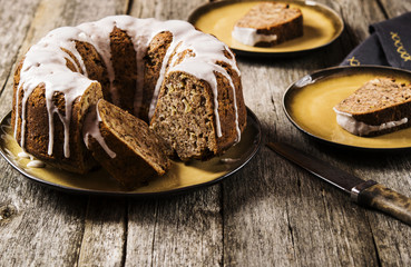 Banana cake with apples and walnuts drizzled frosting and sliced pieces on plates with effect craquelure on the shabby wooden background. Selective focus 