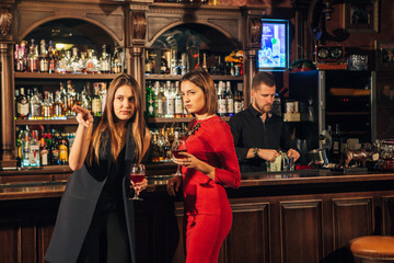Two female friends are in the bar for a drink and talk. The woman with talks near the ear of the...