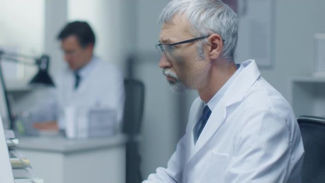 Senior Medical Practitioner Working in His Office at His Desktop Computer. Also His Assistant Works in the Background.  Shot on RED Cinema Camera in 4K (UHD). 
