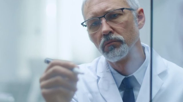 Senior Male Scientific Researcher Writes Scientific Formula on a Glass Whiteboard. He is Wearing Glasses and His Office is Bright and Modern. Slow Motion.  Shot on RED Cinema Camera in 4K (UHD). 
