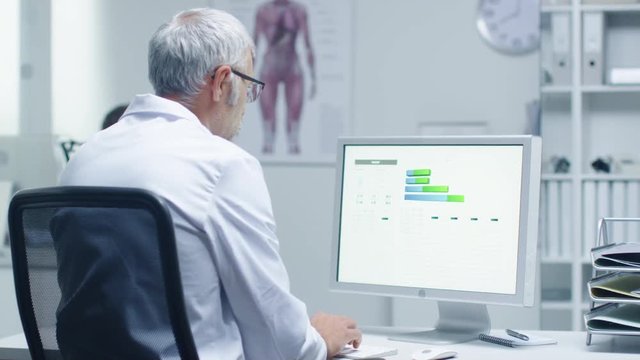 Senior Male Laboratory Researcher Working with Graphs on His Personal Computer. His Assistant Works at His Desk in the Background.  Shot on RED Cinema Camera in 4K (UHD). 