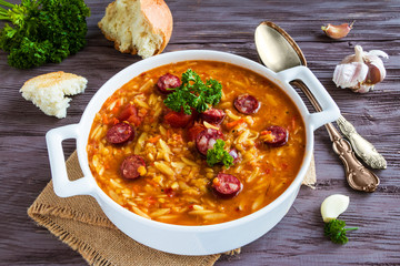 Tomato soup with orzo and smoked sausages in white casserole on wooden rustic table. Fresh bread and parsley, vintage spoon.